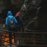person wearing blue hooded jacket standing on footbridge near cave